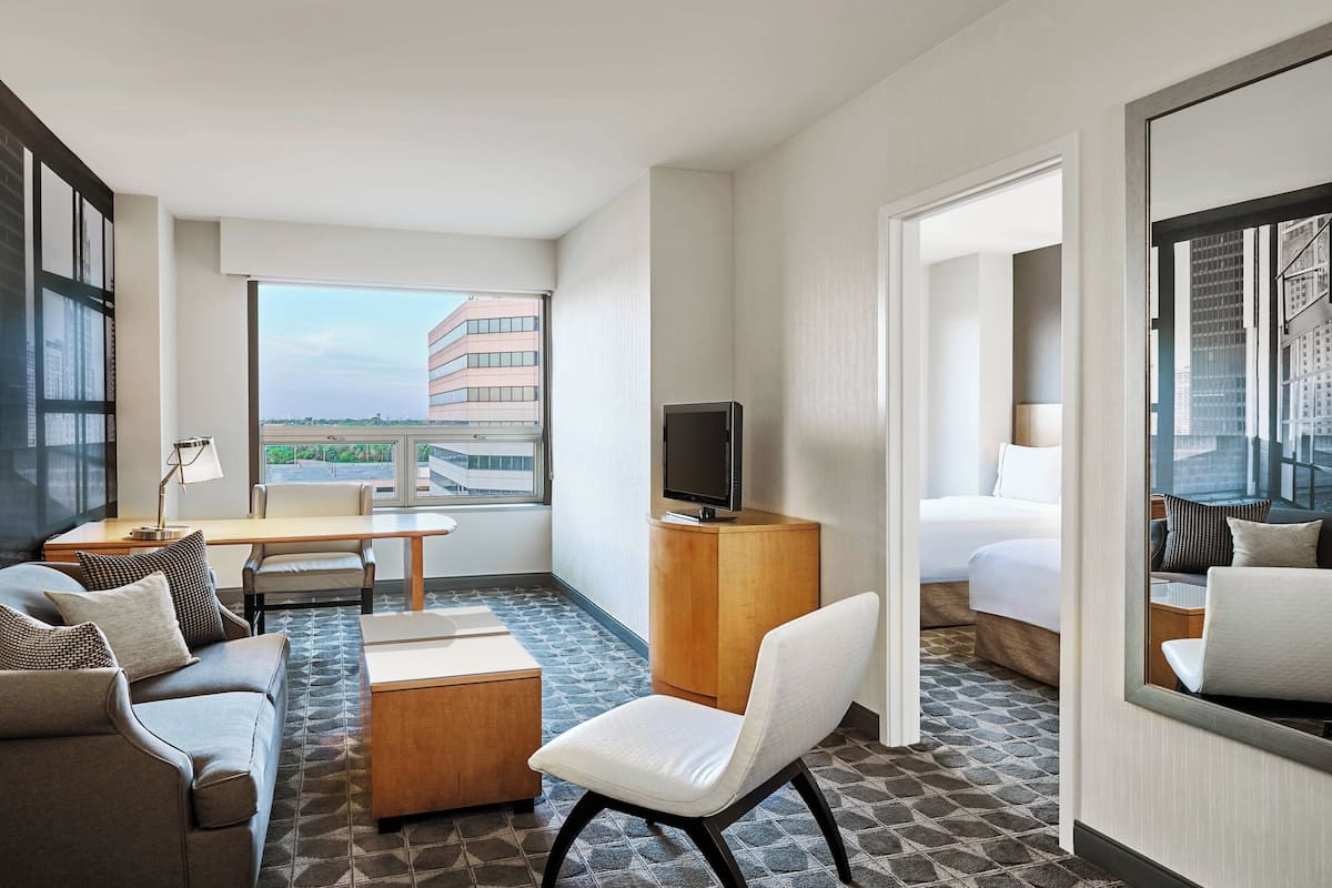 A hotel room with a sofa, a desk, a chair, a bed, a TV, and a large window offering a city view.