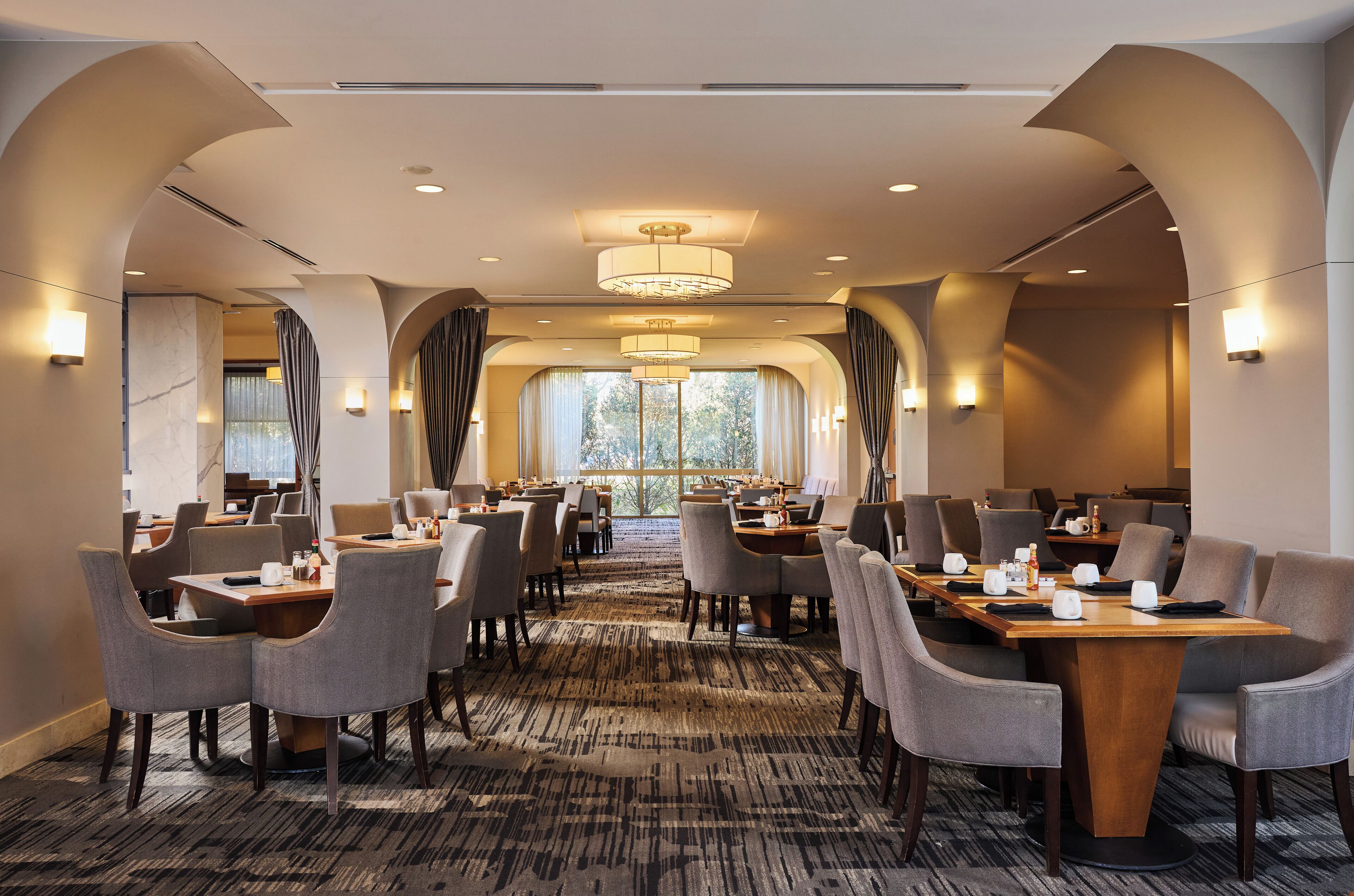 Comedor de hotel con mesas y sillas redondas, ventanas amplias y alfombra estampada.