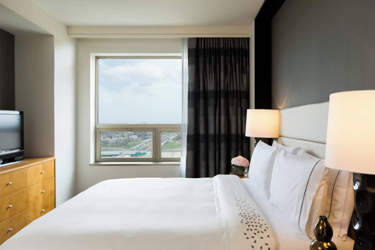 A hotel room with a bed, a TV on a wooden dresser, a window with a view of a cityscape, and a lamp on the nightstand.