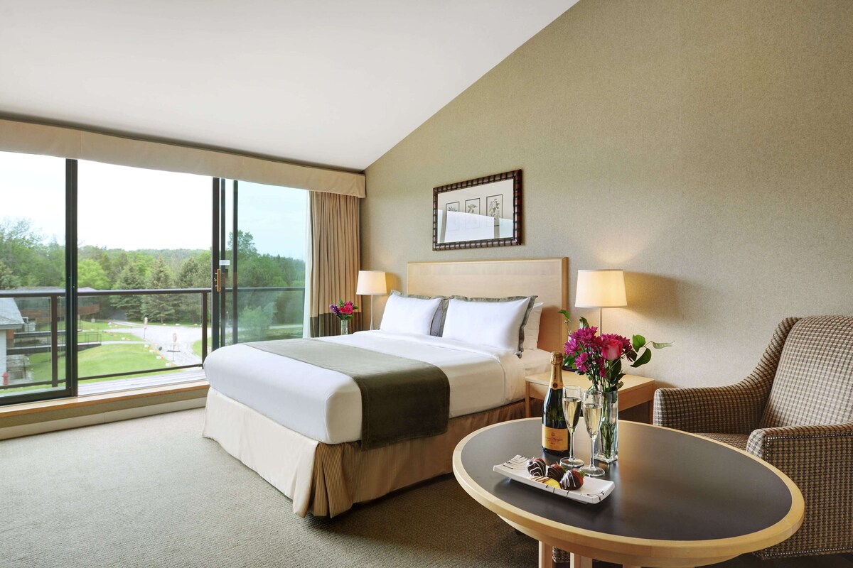 Ein Hotelzimmer mit einem großen Bett, einem runden Tisch mit einer Flasche und Blumen sowie Blick ins Freie.
