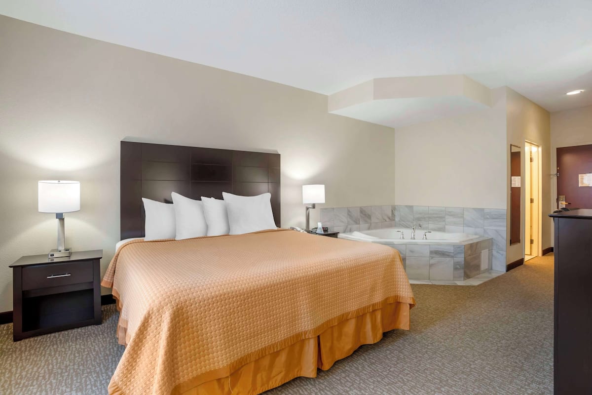 A hotel room with a large bed, two bedside tables with lamps, a built-in headboard, a marble-tiled bathroom with a bathtub, and a dark-colored wardrobe.