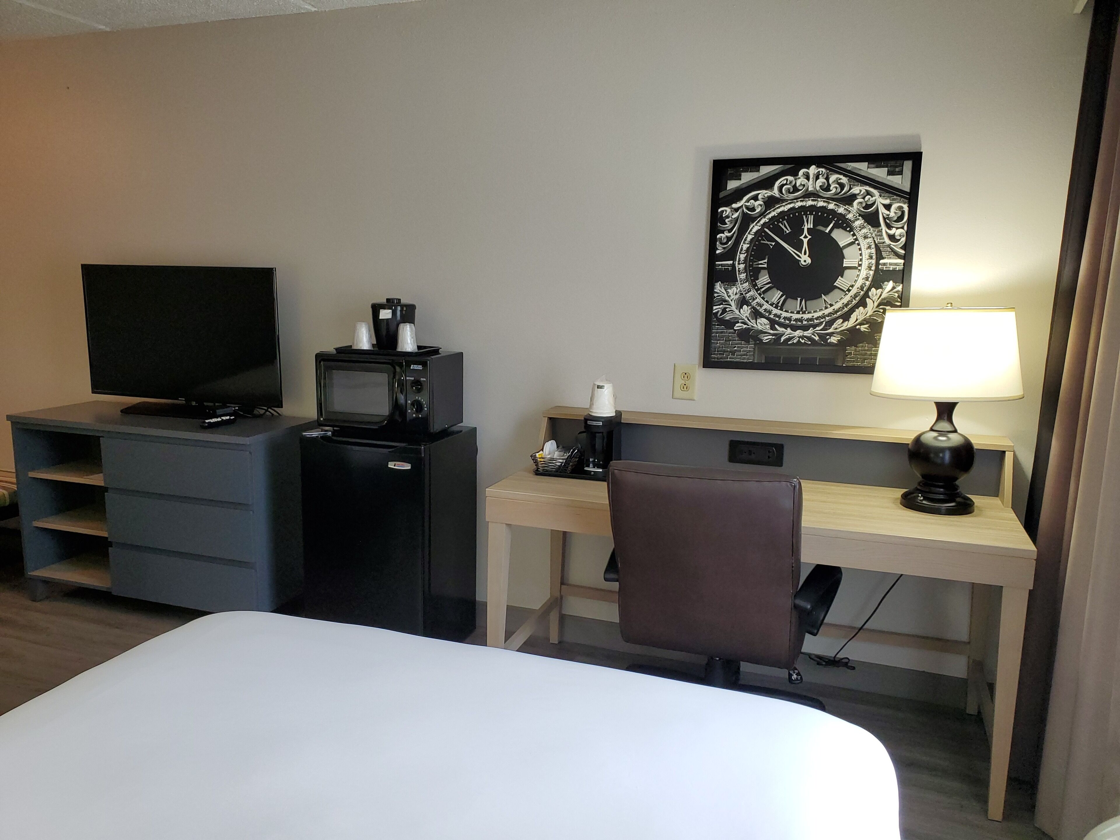 A hotel room with a bed, a desk with a lamp, a chair, a TV, a microwave, and a coffee maker.