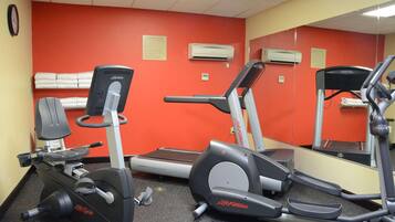Une salle de sport équipée d’appareils de cardio, notamment des tapis de course et un vélo elliptique, et un mur rouge orné d’une horloge et d’un panneau indiquant les règles à respecter pour faire de l’exercice.