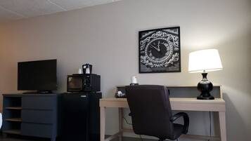 A hotel room with a desk, a television, a microwave, a coffee maker, a chair, and a clock on the wall.