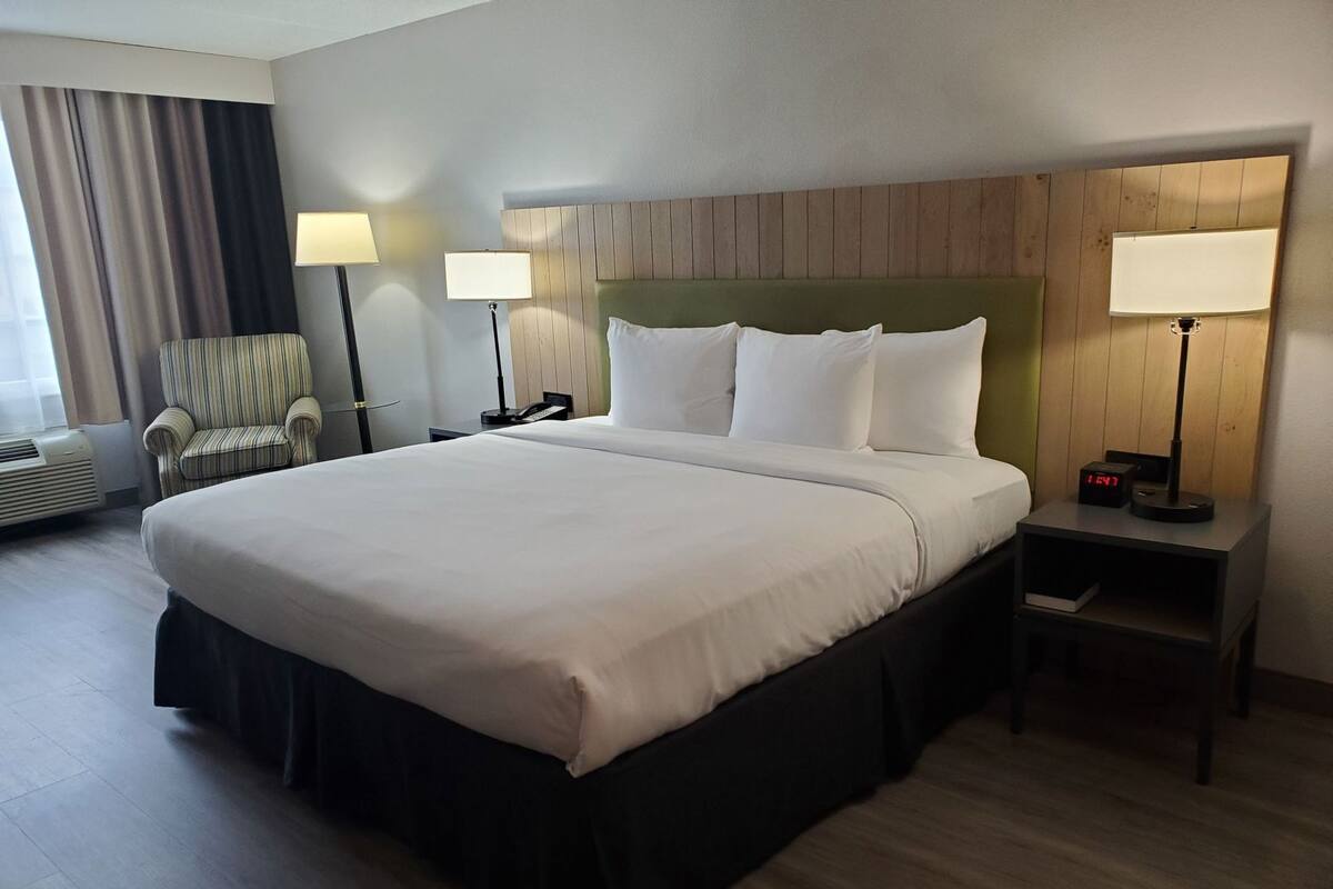 A hotel room with a large bed, two bedside lamps, a chair, a nightstand with a clock, and a window with curtains.