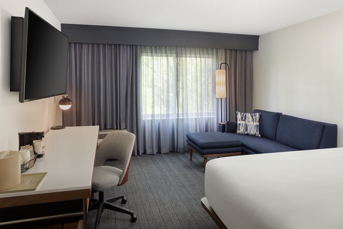 Une chambre d’hôtel comprenant un lit, un canapé, un bureau avec une chaise, un téléviseur fixé au mur et une grande fenêtre avec des rideaux.