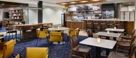 A modern restaurant with a bar area, multiple seating options including round tables and bar stools, and a visible kitchen area.