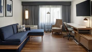 A modern hotel room with a sofa, armchair, desk, and TV.