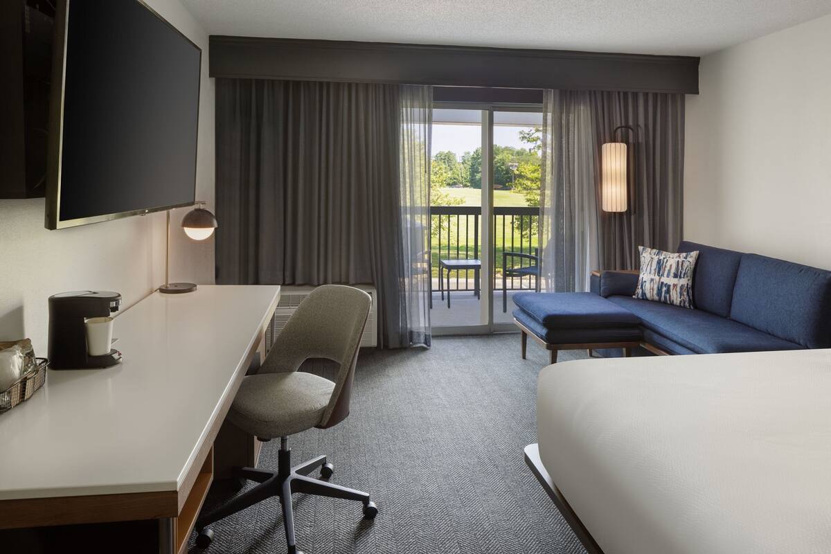 Une chambre d’hôtel comprenant un lit, un bureau avec une chaise, un canapé, un téléviseur fixé au mur et un balcon avec des rideaux.