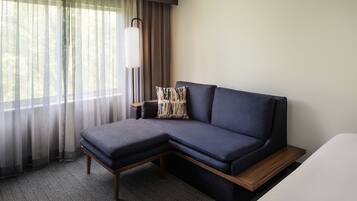 A modern hotel room with a sectional sofa, a wooden bench, a large window with sheer curtains, and a floor lamp.