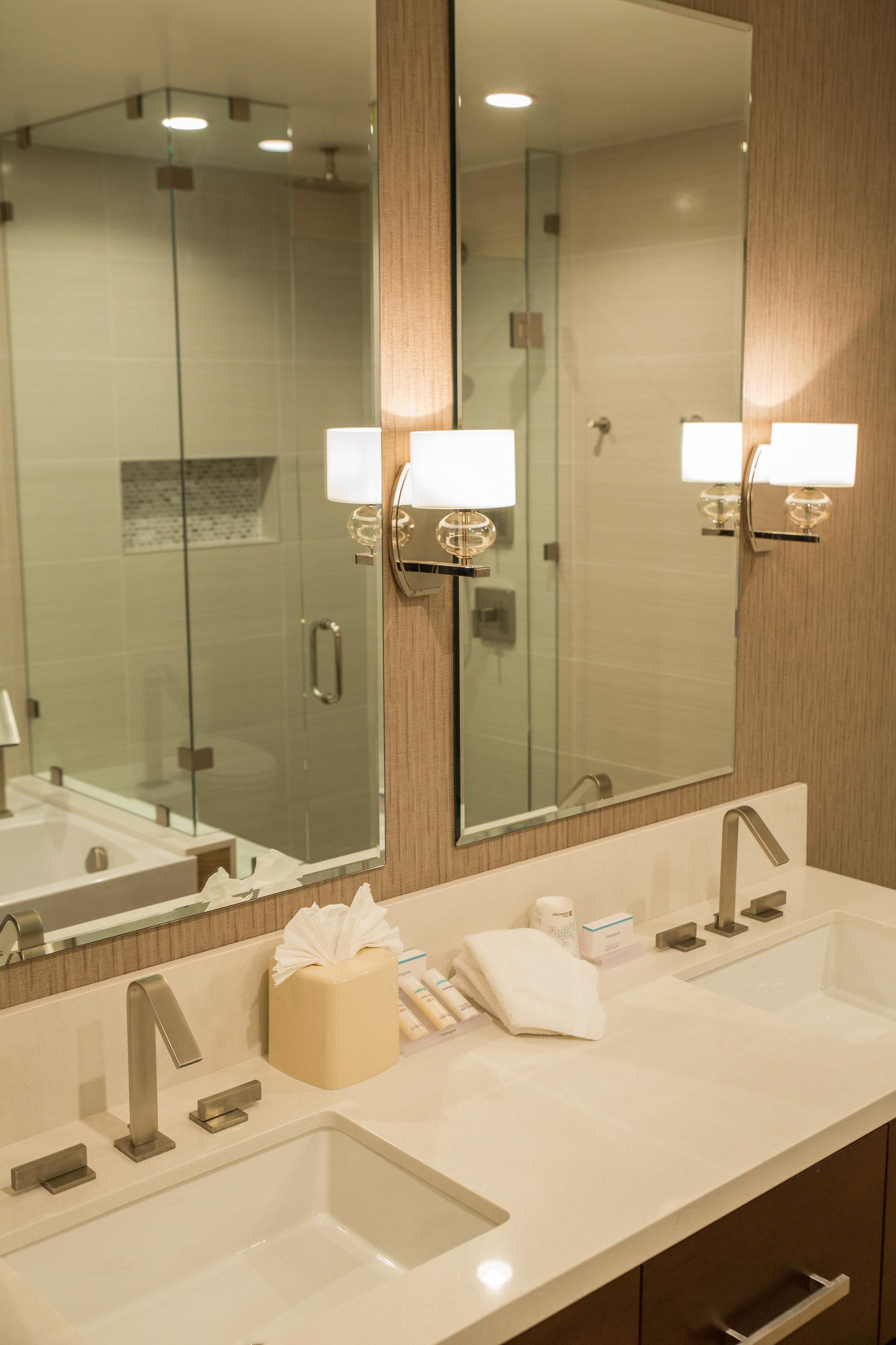 A hotel bathroom with a large mirror, two sinks, a towel dispenser, and toiletries.