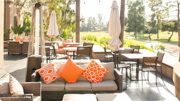 Un espacio al aire libre con mobiliario de mimbre, cojines naranjas y luces colgantes.