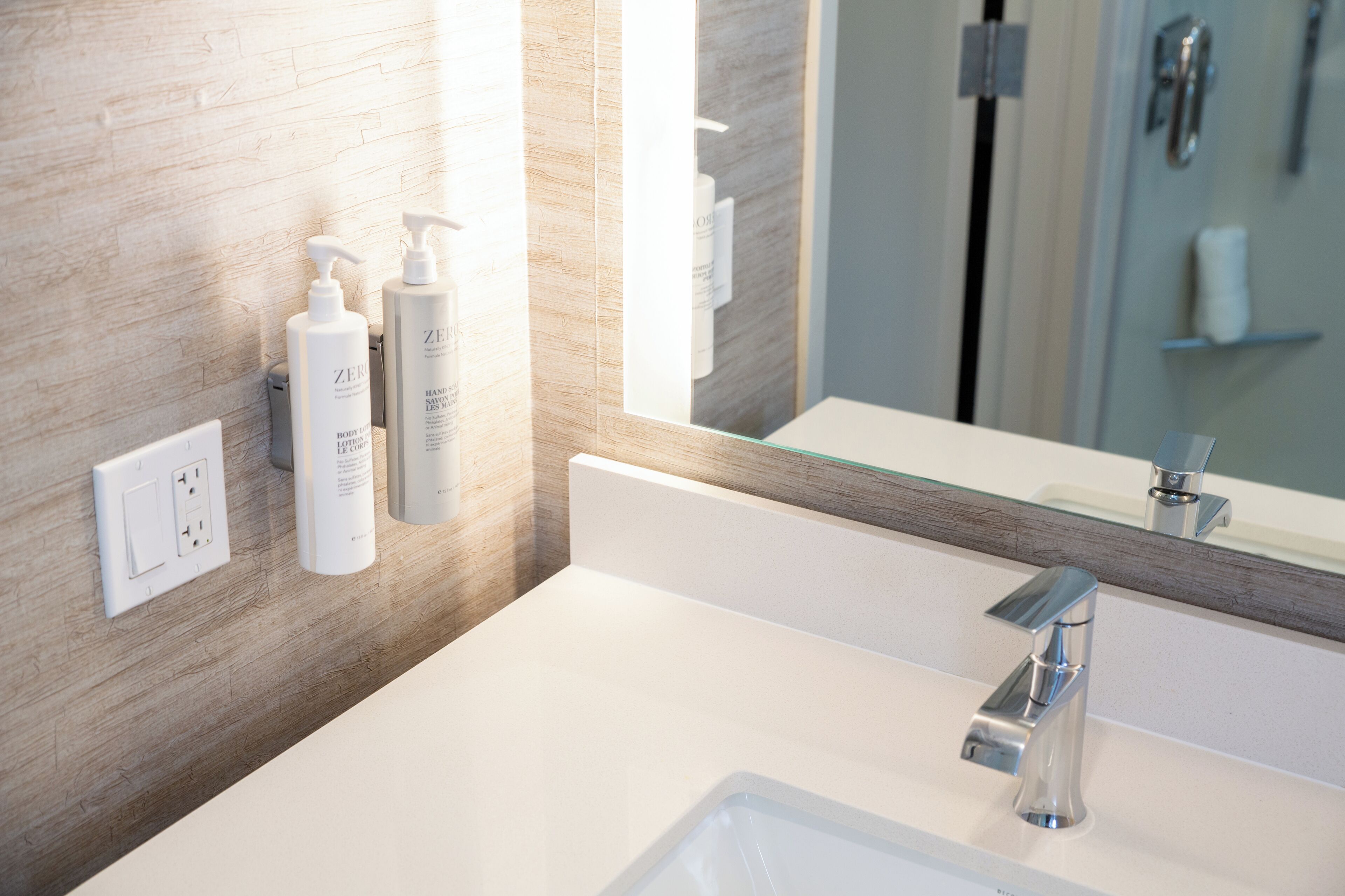 A modern bathroom with a white sink, a chrome faucet, and a mirror above it. There are two bottles of hand soap on the wall.
