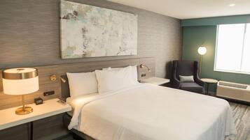 A hotel room with a bed, bedside table, lamp, chair, and artwork on the wall.