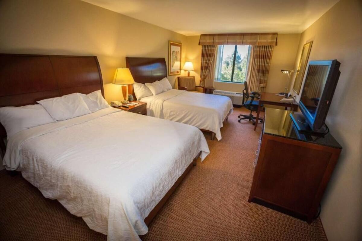 A hotel room with two beds, a desk, a chair, a TV, and a window with curtains.