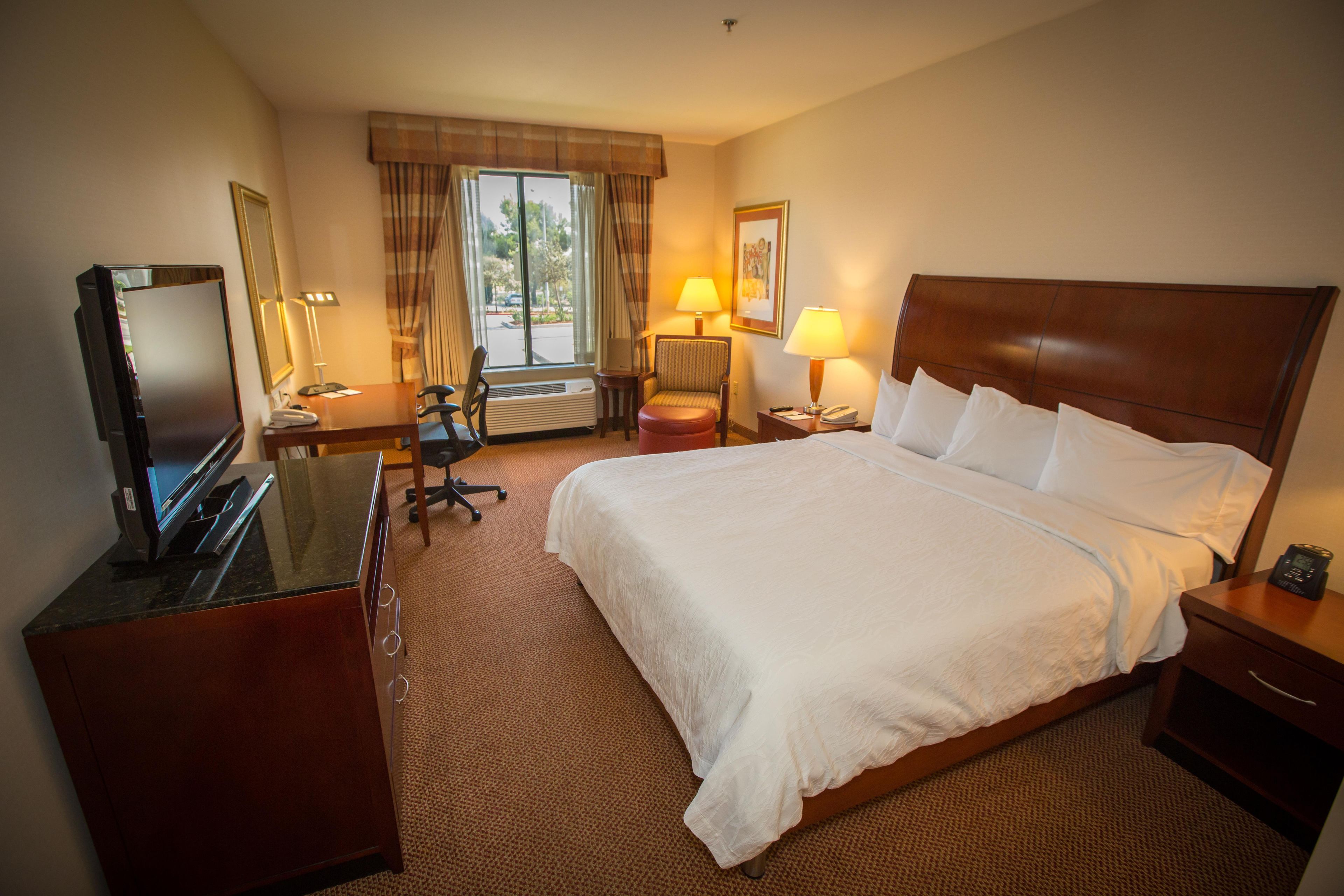 A hotel room with a bed, a desk with a computer, a chair, a lamp, and a window with curtains.