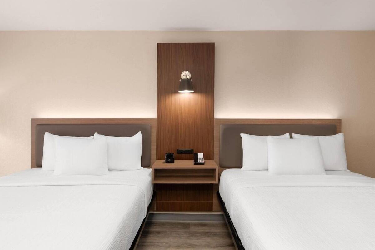 Quarto de hotel com duas camas, cabeceira de madeira, mesa de cabeceira com abajur e uma pequena mesa.