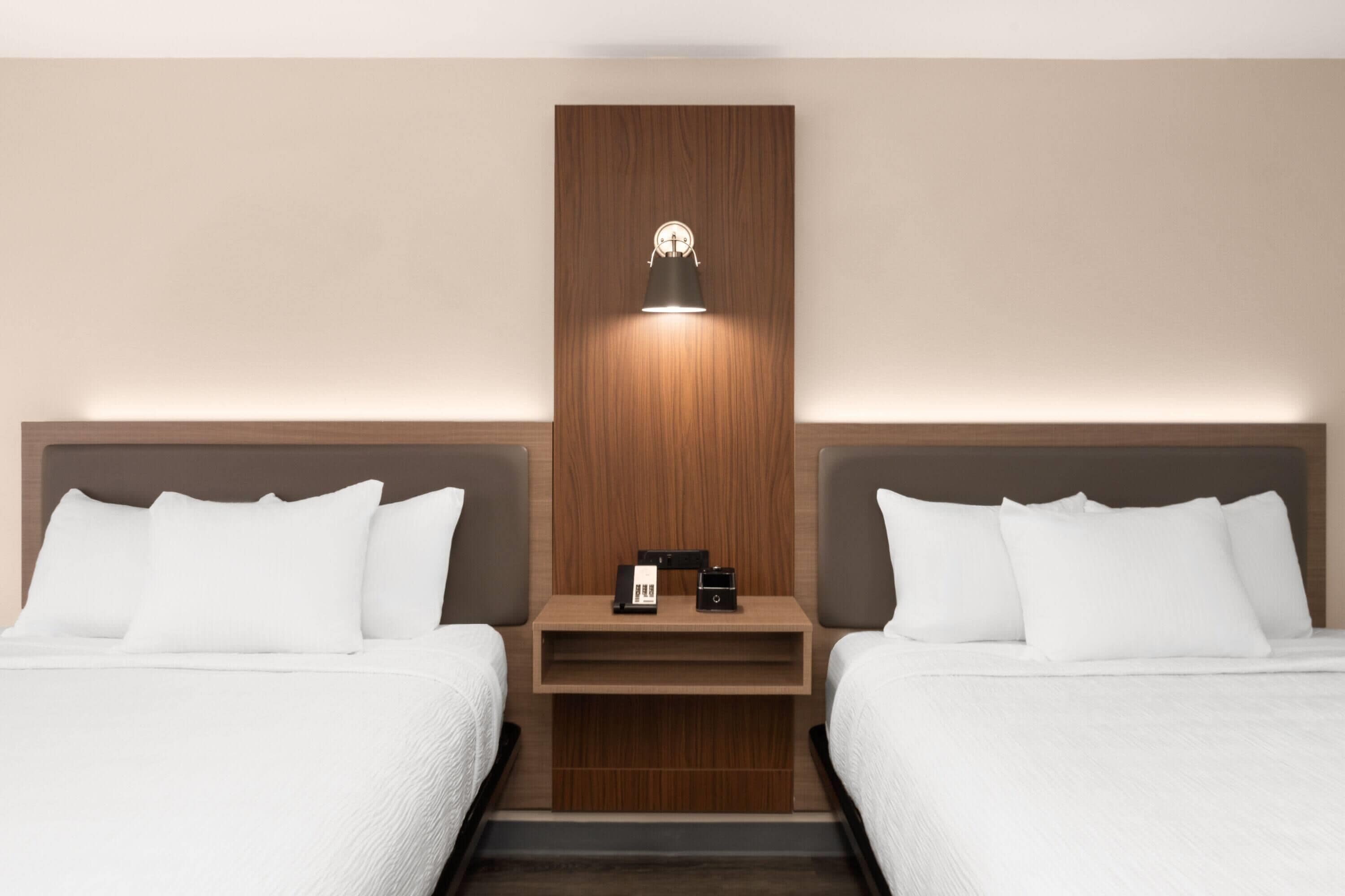 A hotel room with two beds, a wooden panel wall, a nightstand with a lamp, and a small table with a phone and charger.