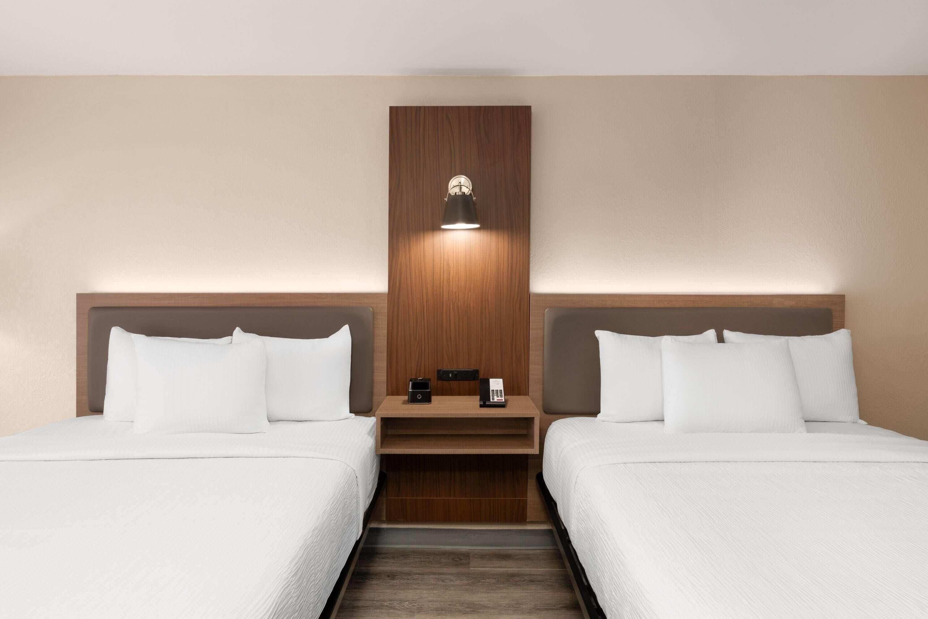 A hotel room with two beds, a wooden headboard, a nightstand, and a wall-mounted lamp.