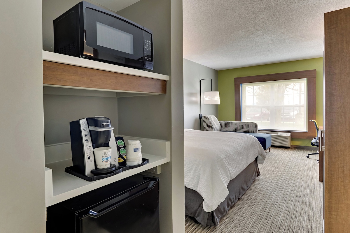 A hotel room with a kitchenette, a bed, a window, and a lamp.