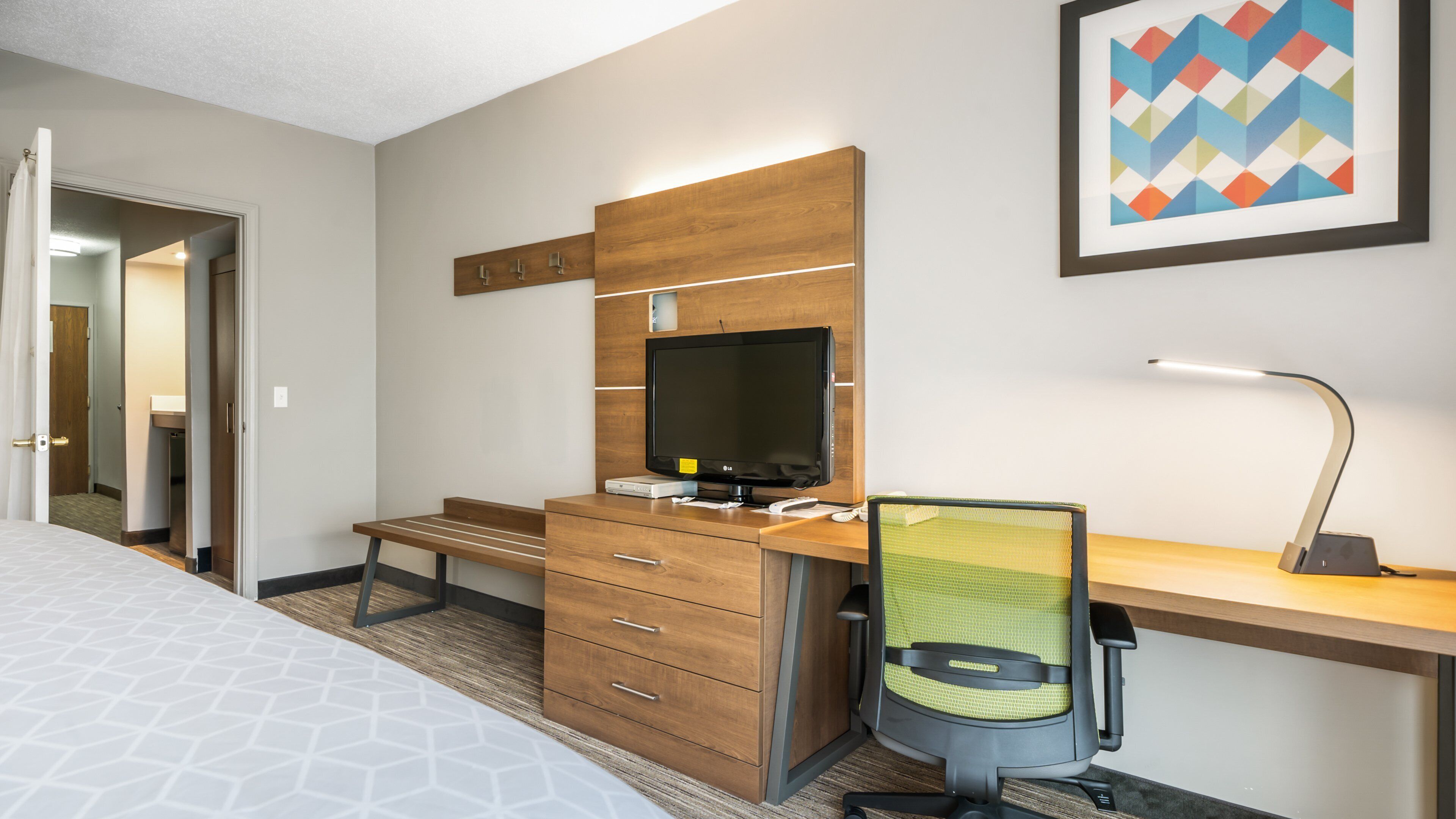 Une chambre d’hôtel avec un lit, un bureau en bois avec un ordinateur, une chaise, une lampe de bureau et un tableau encadré au mur.