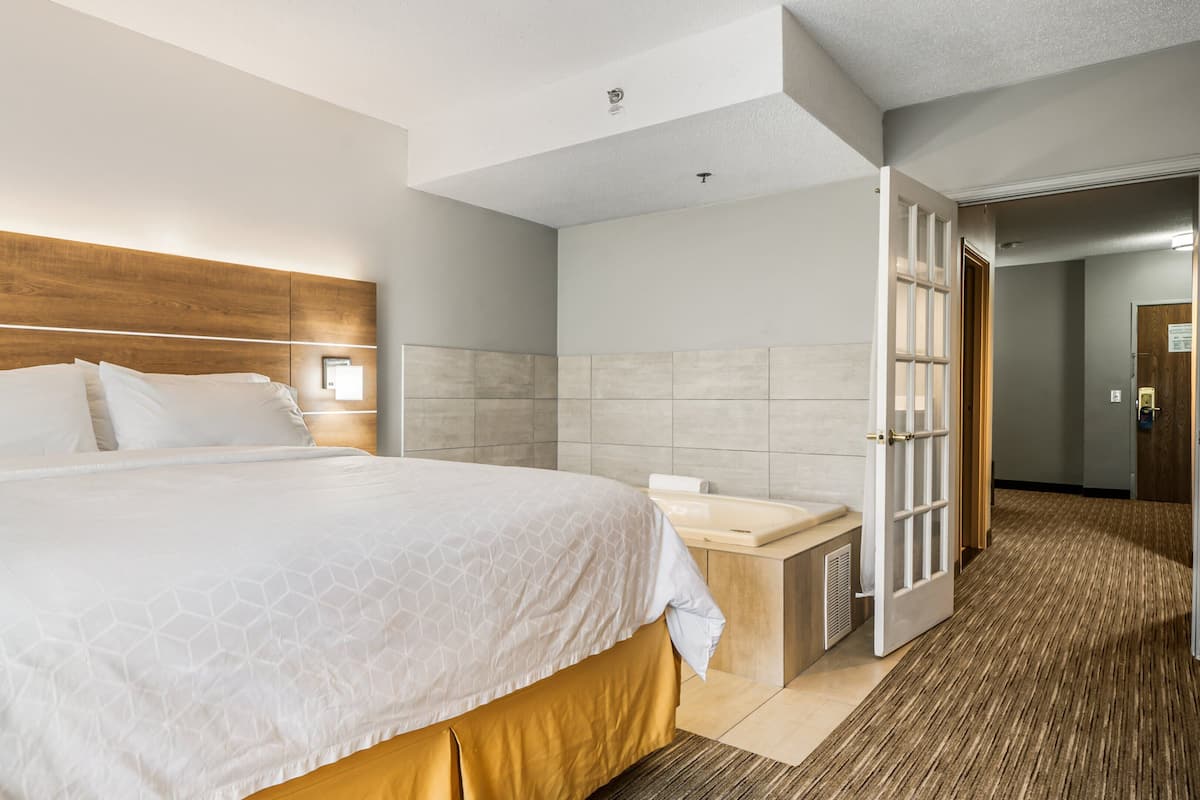 A hotel room with a large bed, a wooden headboard, a bedside table with a lamp, and a bathroom with a shower and a toilet.