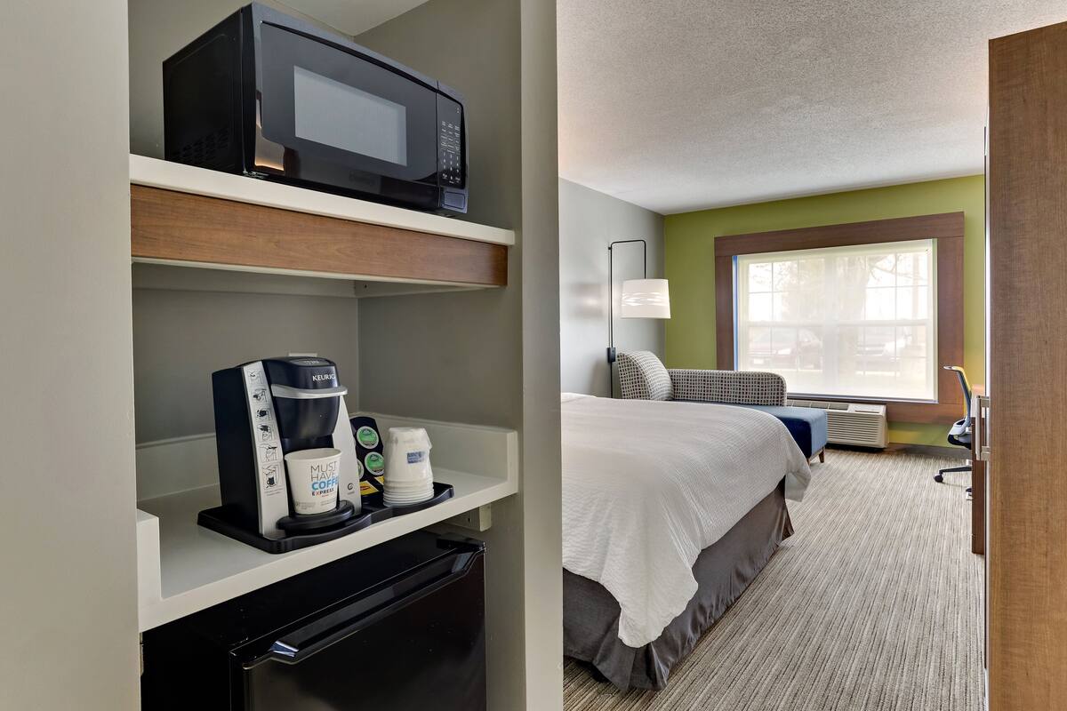 A hotel room with a kitchenette, a bed, a window, and a lamp.