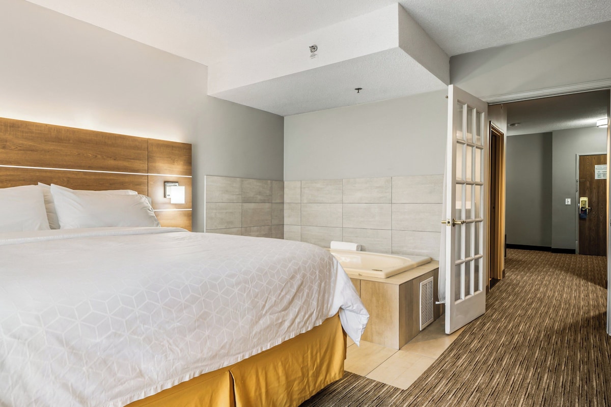 A hotel room with a large bed, a wooden headboard, a bedside table with a lamp, and a bathroom with a shower and a toilet.