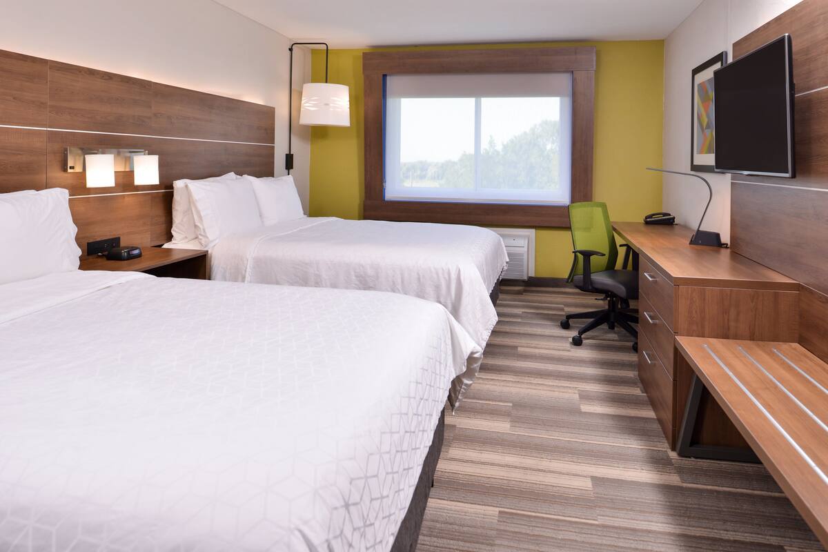 A hotel room with two beds, a desk, a chair, a flat-screen TV, and a window with a view of trees.