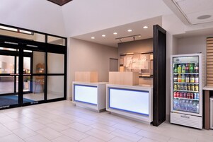 Un vestíbulo de hotel con una recepción, una máquina expendedora de bebidas y una entrada por puerta de cristal.