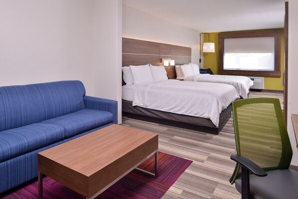 A hotel room with a blue sofa, a wooden coffee table, a green chair, two beds with white bedding, a large window with blinds, and a wall-mounted lamp. - Holiday Inn Express Hotel & Suites Elgin by IHG (Elgin)