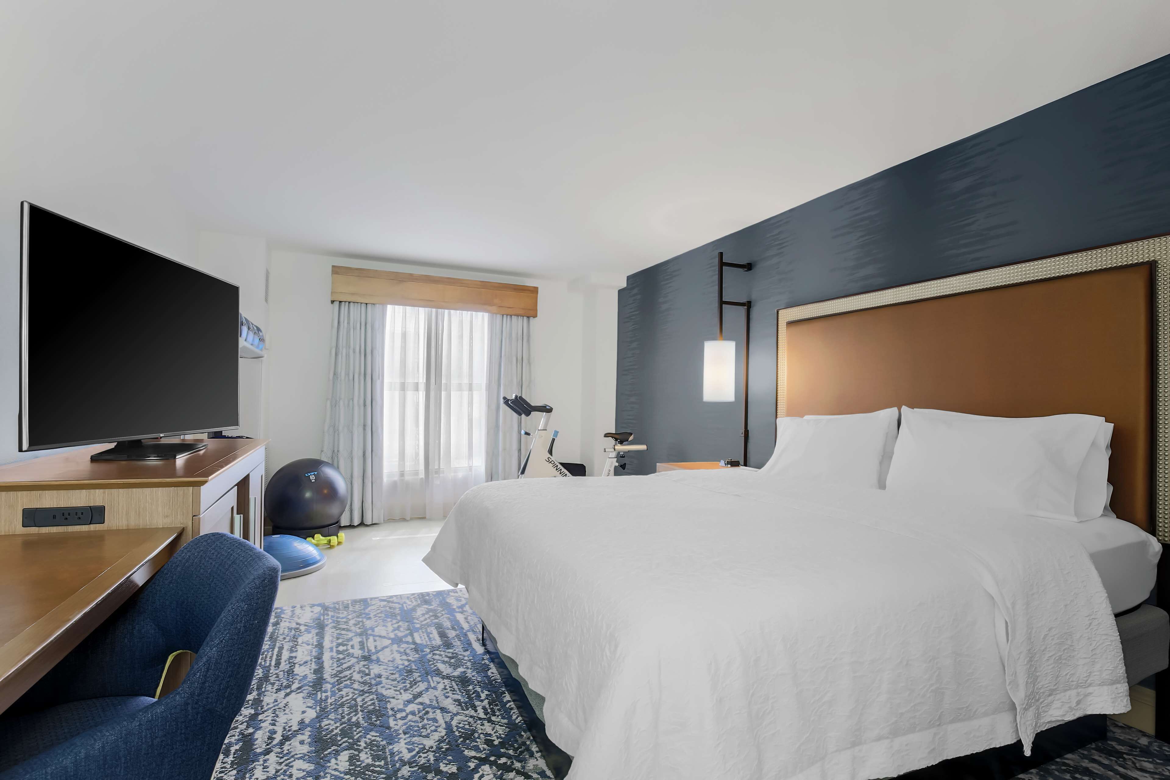 Une chambre d’hôtel avec un grand lit, un bureau doté d’un téléviseur à écran plat, du matériel d’exercice et une fenêtre avec des rideaux.