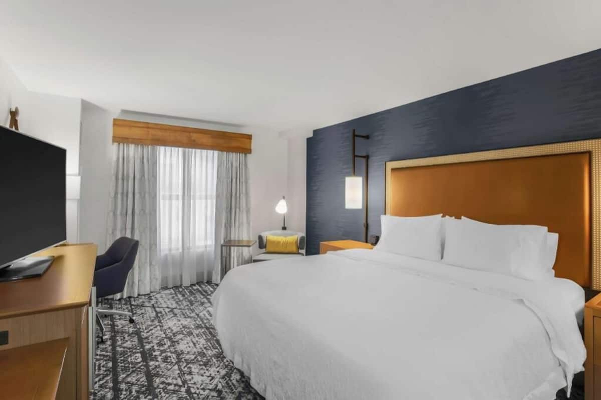 A hotel room with a large bed, a desk, a chair, a TV, a lamp, and a window with curtains.
