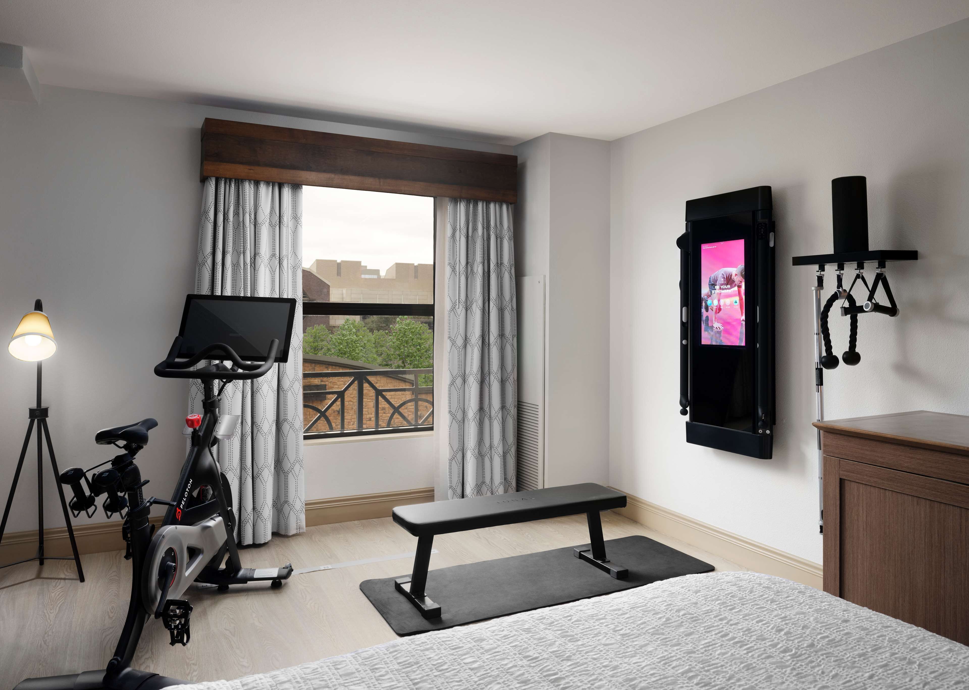 Ein Hotelzimmer mit Bett, Ergometer, Sitzbank, Fernseher und Blick ins Freie.