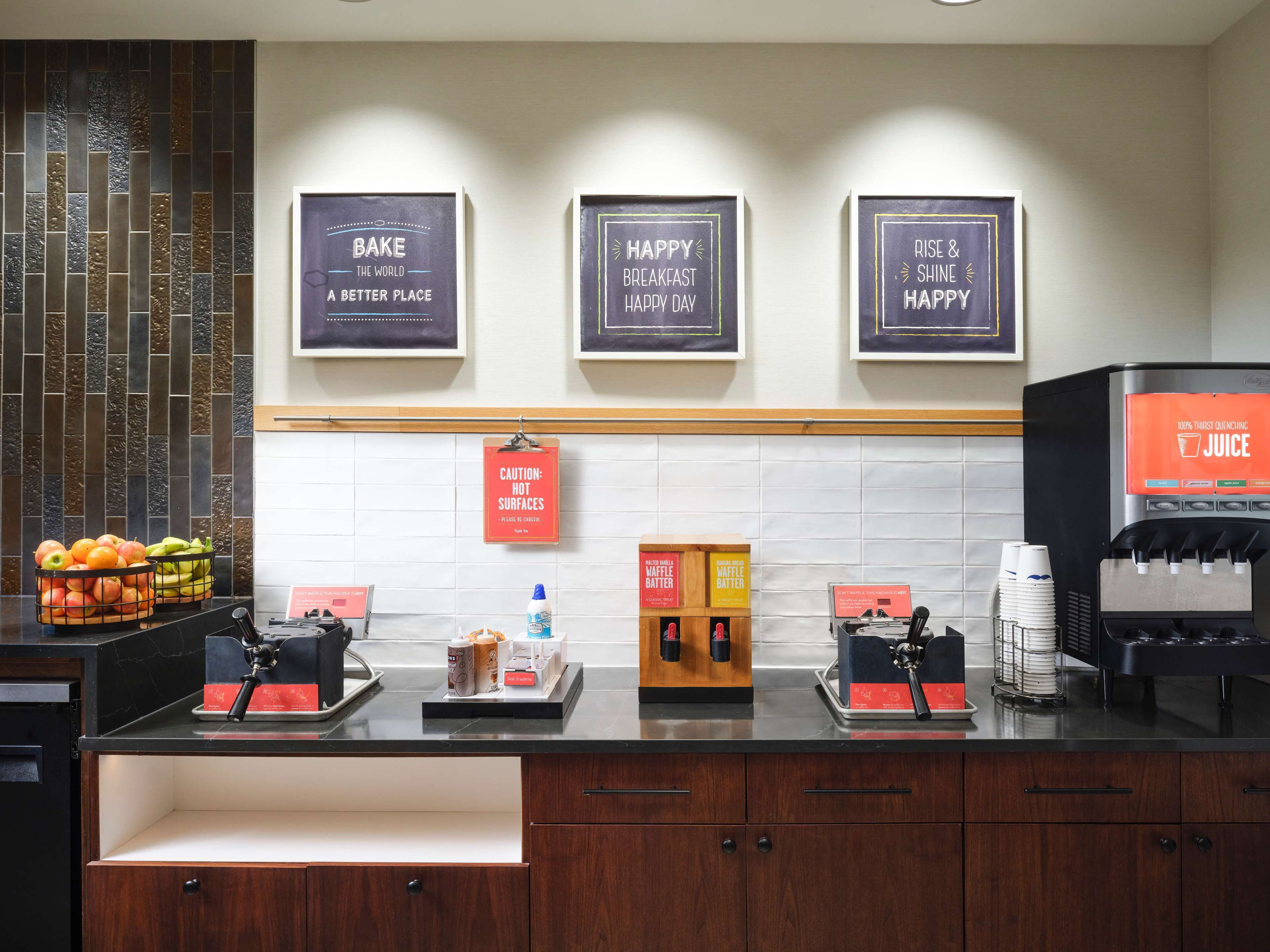 Un banco della colazione con macchina del caffè, piastra per waffle e distributore di succhi di frutta. Ci sono cartelli appesi al muro e un cartello di avvertimento per superfici calde.