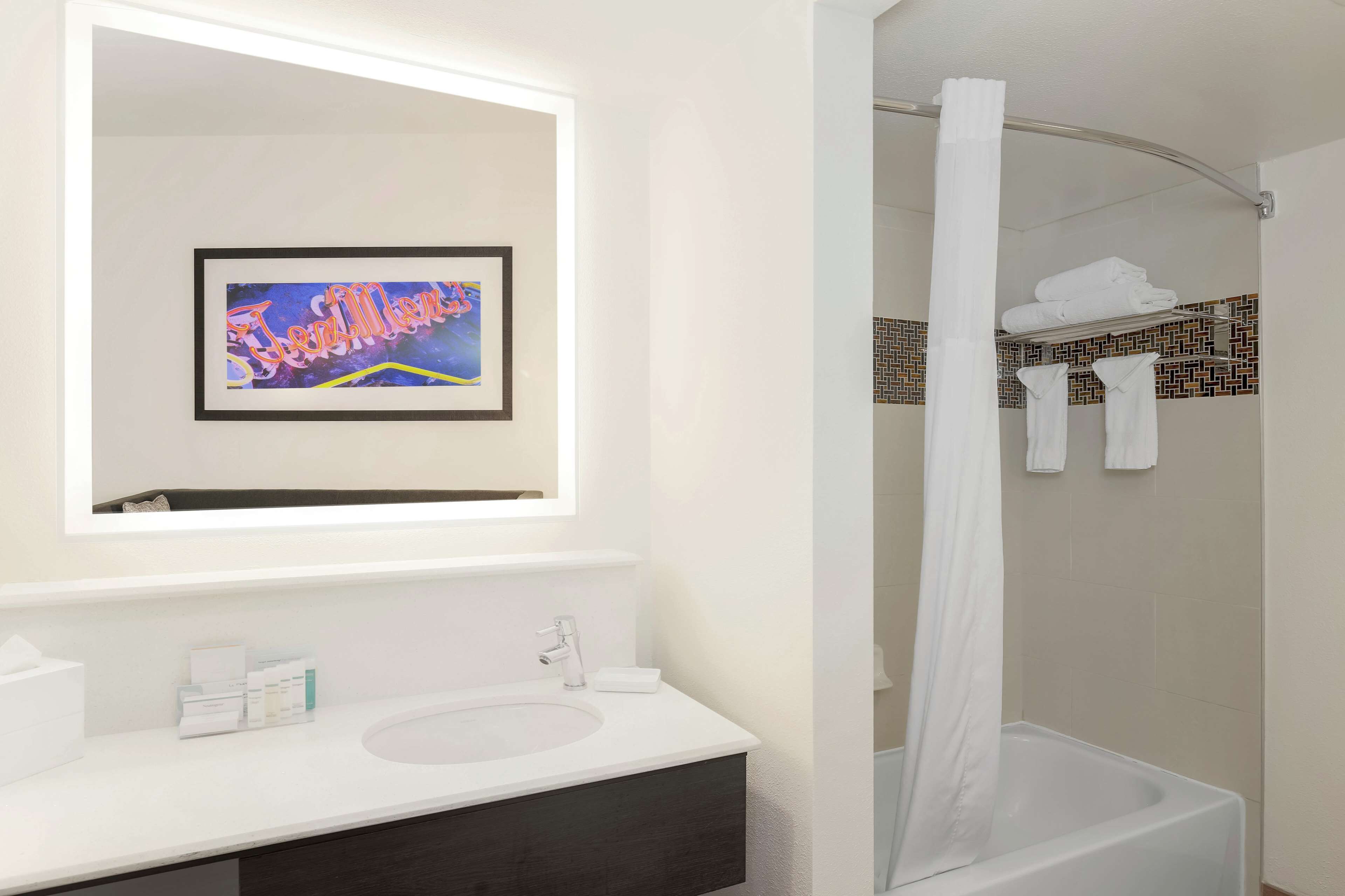 Un baño moderno con un tocador blanco, una obra de arte enmarcada sobre el espejo y una ducha con cortina blanca.