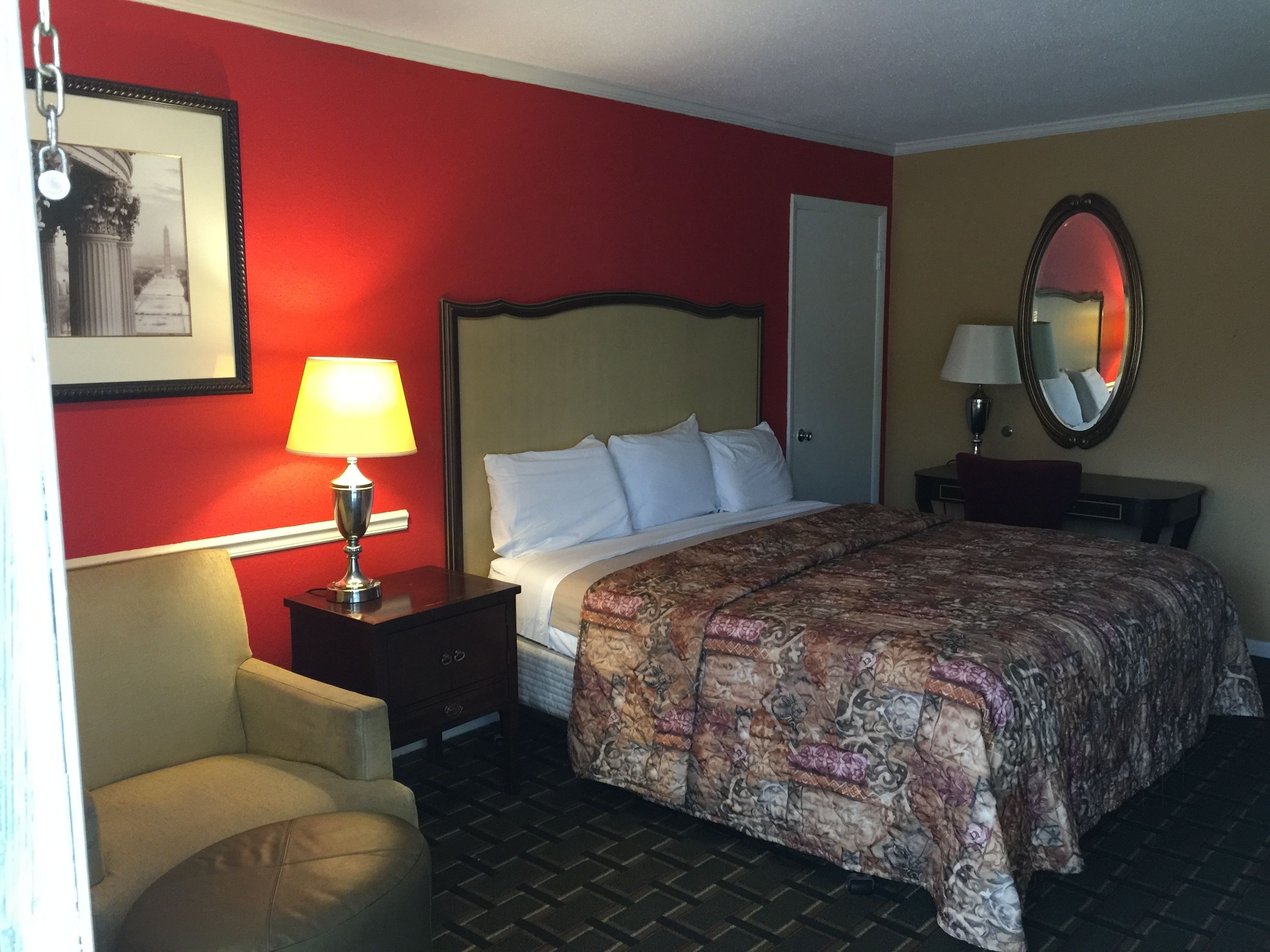 A hotel room with a bed, a chair, a nightstand with a lamp, a dresser, a door, and a mirror on the wall.