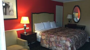 A hotel room with a bed, a chair, a nightstand with a lamp, a dresser, a door, and a mirror on the wall.