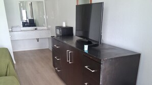 Ein Schlafzimmer mit Holzboden, einer grünen Bettdecke, einer dunklen Holzkommode mit einem Flachbildfernseher, einem kleinen Hocker und einem Spiegel über der Kommode.
