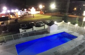 A swimming pool with a blue tint, surrounded by a white fence and a parking area with cars.