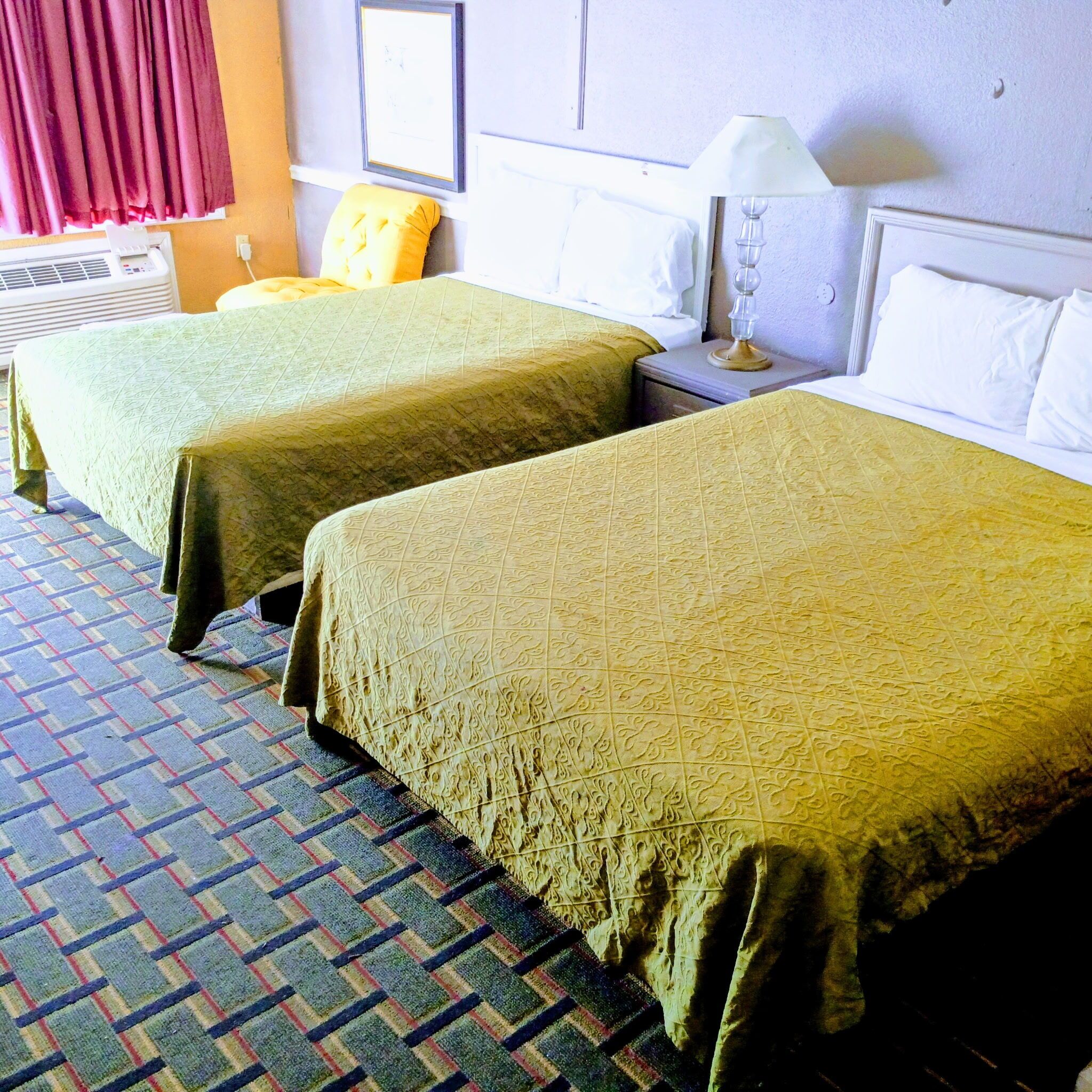A hotel room with two beds, a patterned carpet, a window with curtains, a wall-mounted air conditioner, and a bedside lamp.