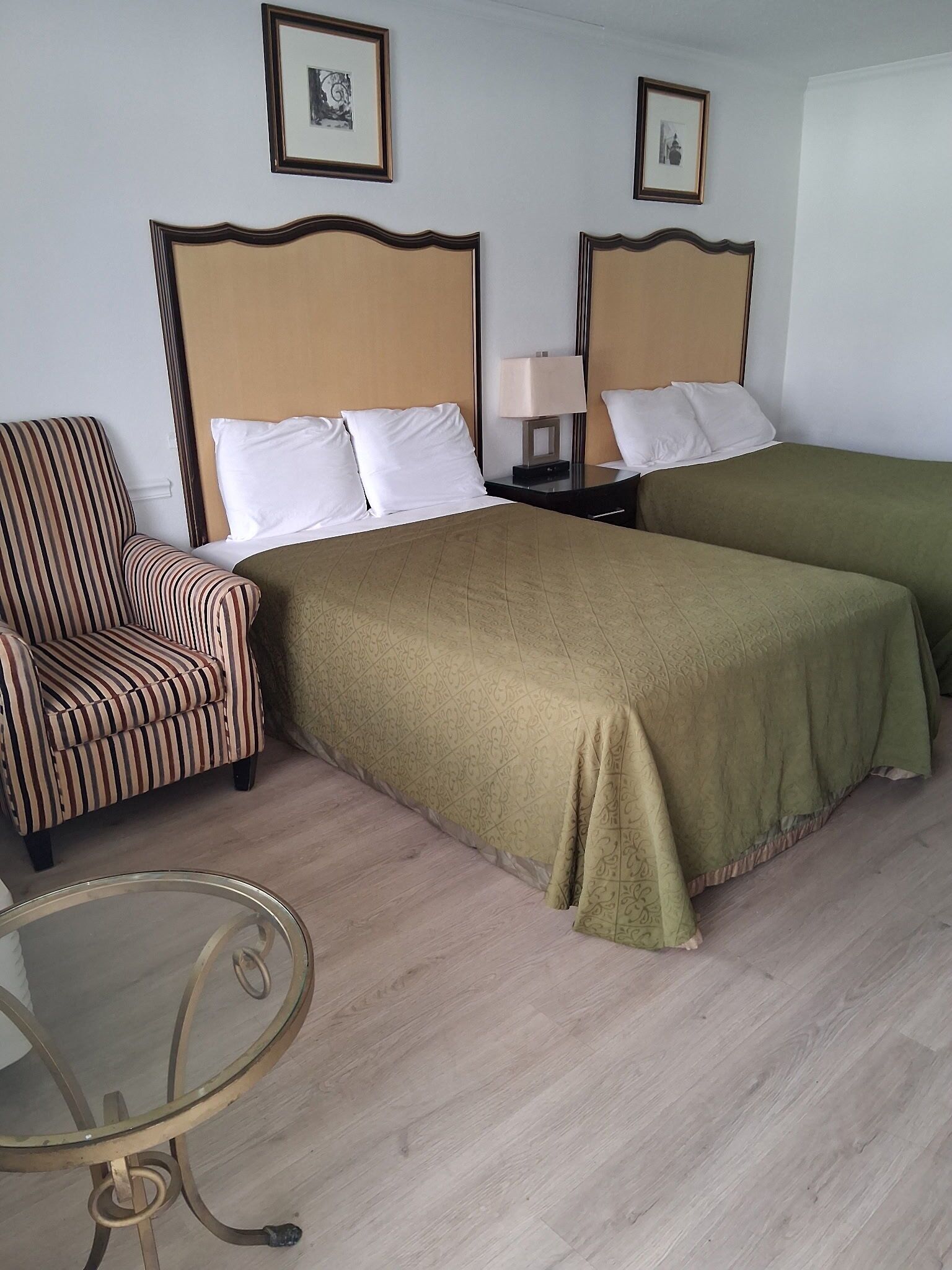 A hotel room with two single beds, a striped armchair, a bedside table with a lamp, and a glass table.
