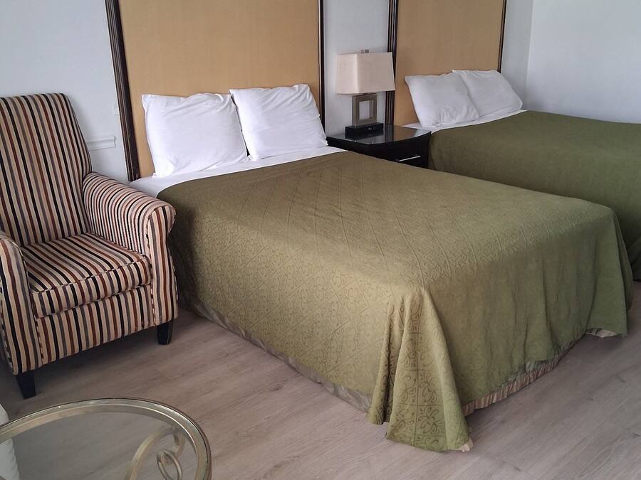 A hotel room with two single beds, a striped armchair, a bedside table with a lamp, and a glass table.