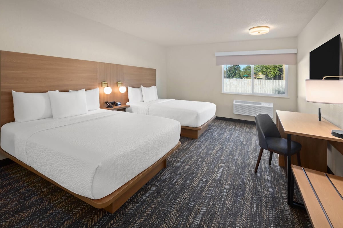A hotel room with two beds, a desk, a chair, a TV, and a window with a view of trees.