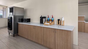 Cozinha moderna com geladeira de aço inoxidável, micro-ondas e armários de madeira. Há duas embalagens de sabonete líquido e uma cafeteira sobre a bancada.
