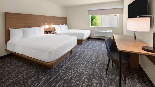 Quarto de hotel com duas camas, uma escrivaninha, uma cadeira, um abajur, uma televisão e uma janela com vista para árvores.