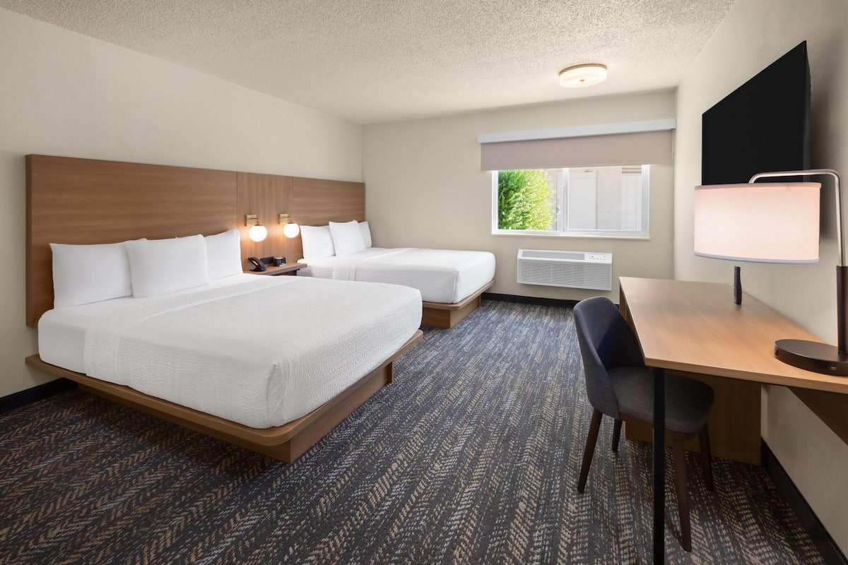 A hotel room with two beds, a desk, a chair, a lamp, a TV, and a window with a view of trees.