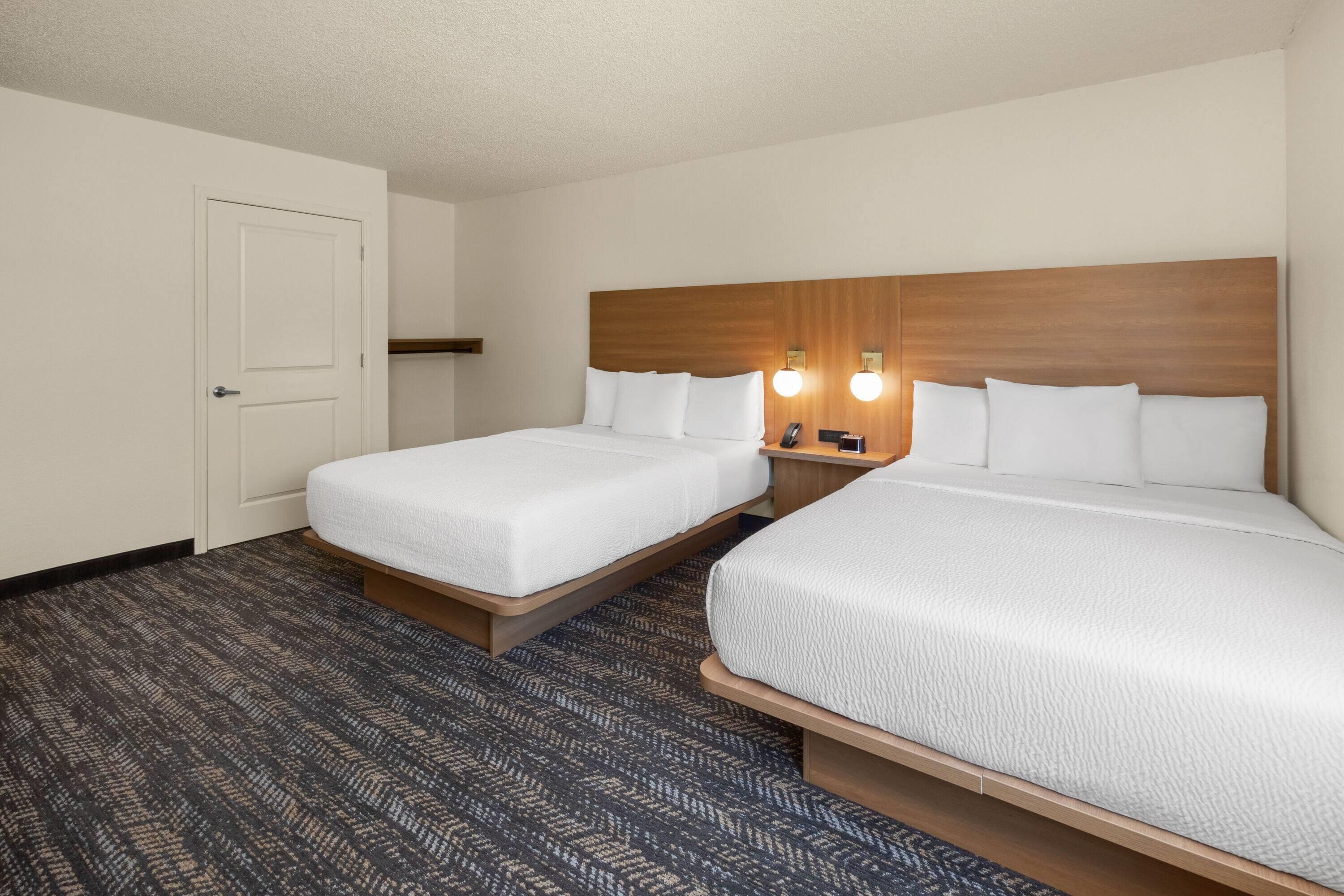 A hotel room with two beds, a wooden headboard, a bedside table with a lamp, and a patterned carpet.
