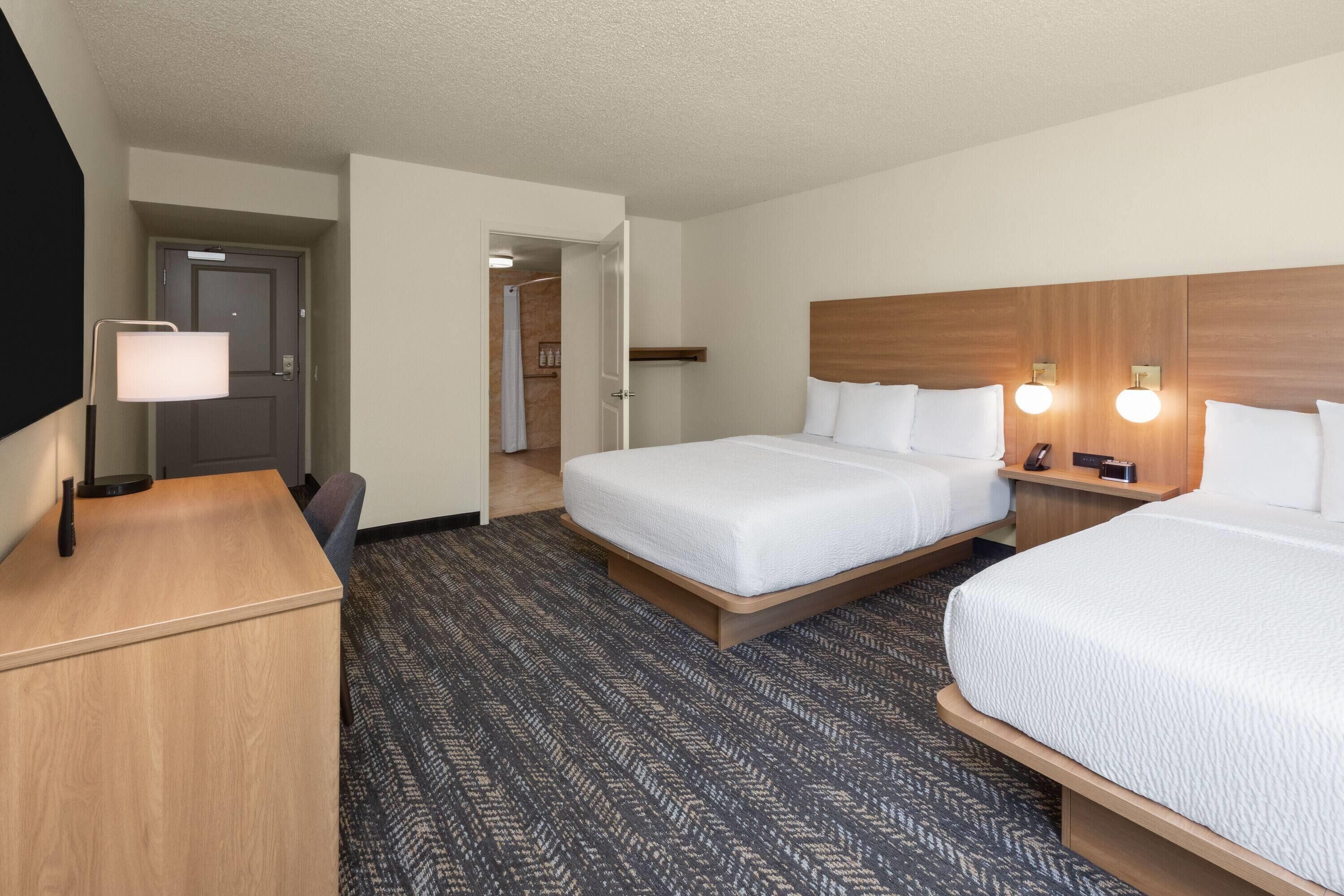 A hotel room with two beds, a desk, a lamp, and a shower area.