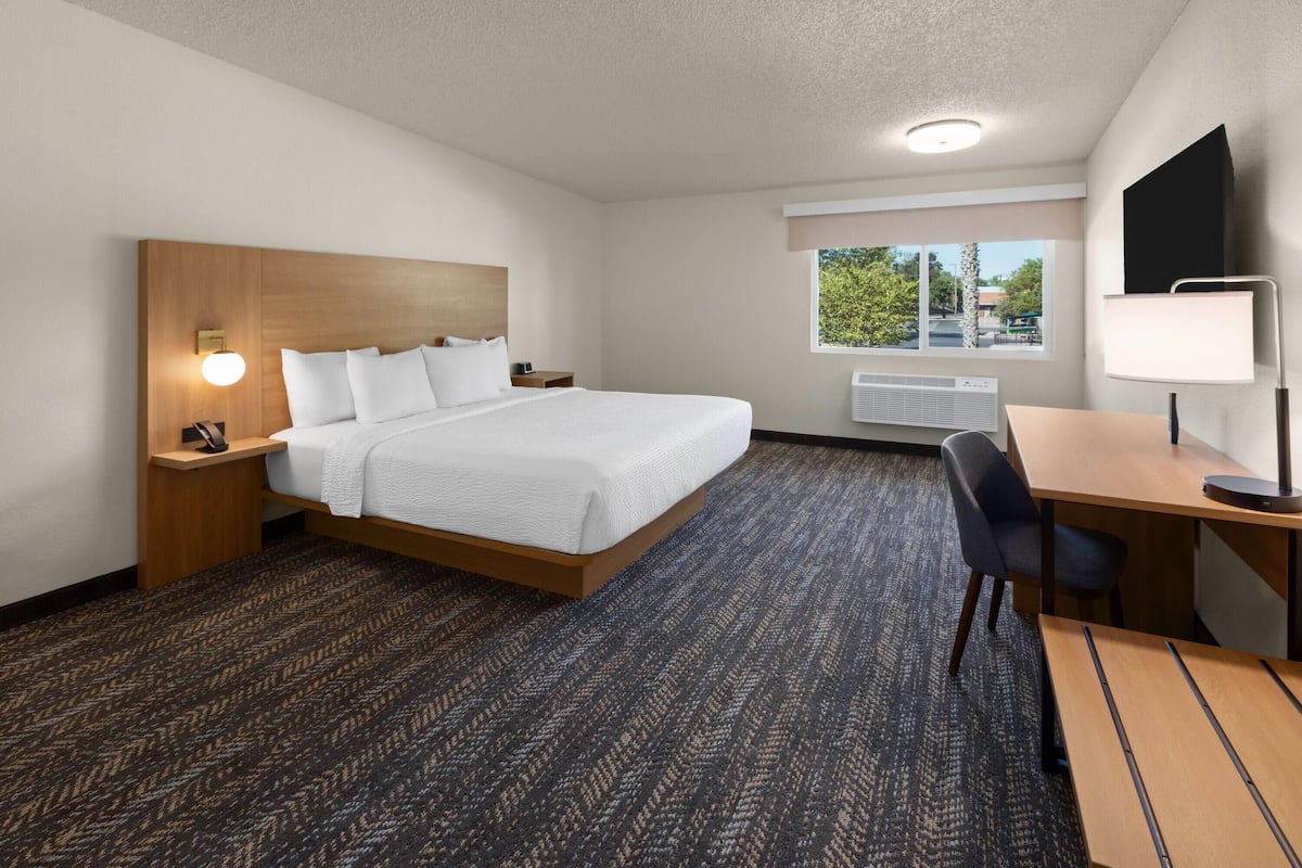 A hotel room with a large bed, a desk, a chair, a nightstand, a wall-mounted lamp, and a window with a view of the outside.
